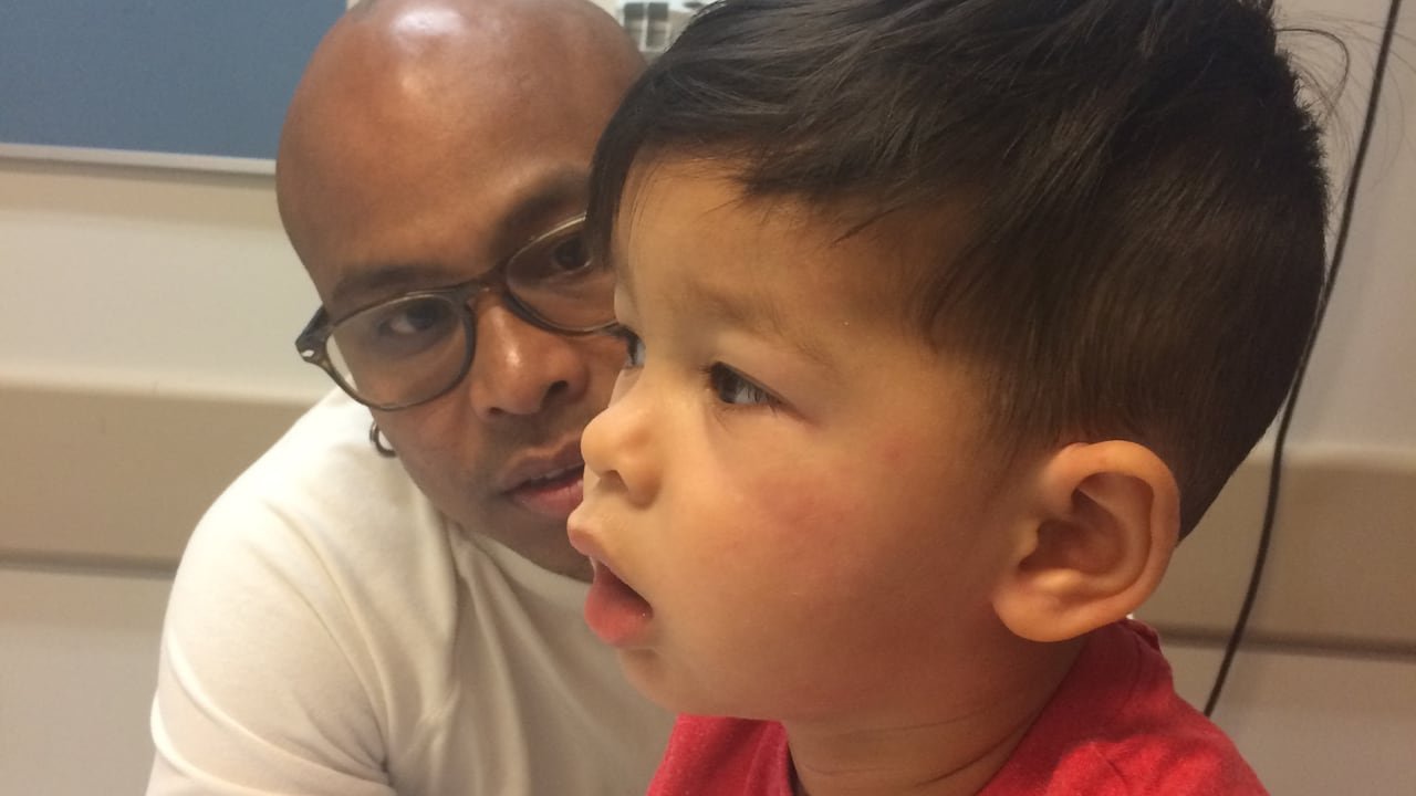 default-168 In a hospital room, a man looks concerned at a boy breathing with his mouth open and who has hives on his cheeks.