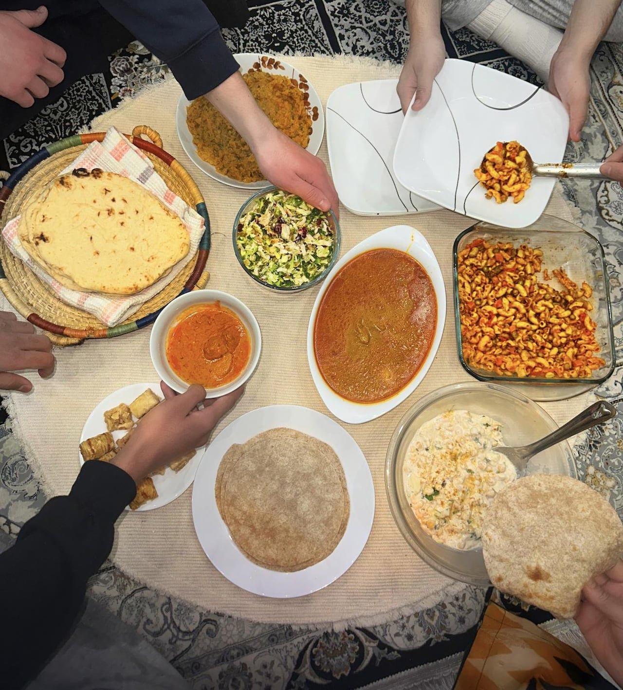 default-239 A bird’s eye view of several hands reaching for food, such as butter chicken, macaroni salad, roti and baklava.