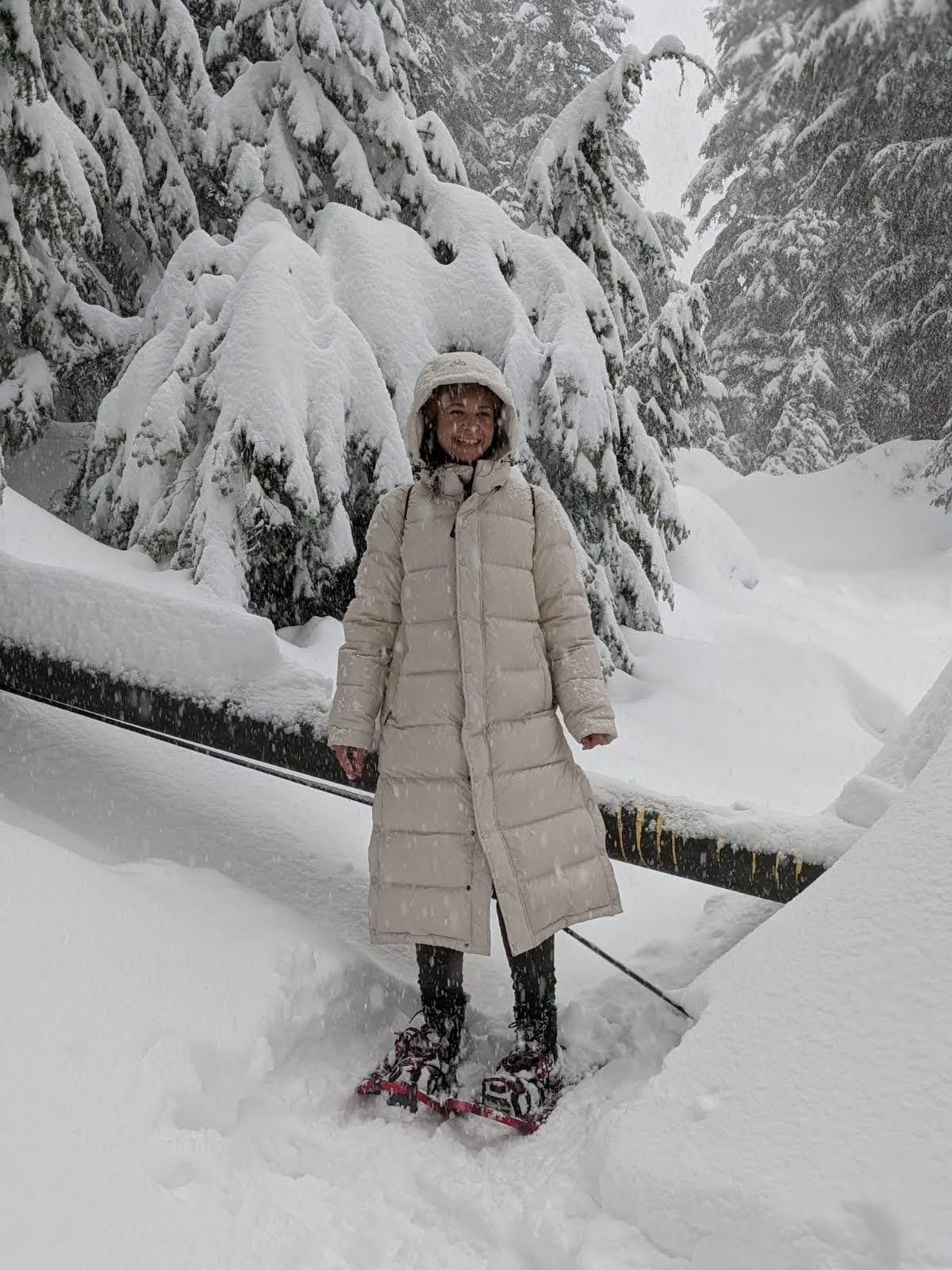 default-25 A woman in a white poofy jacket and snowshoes smiles as she poses on a snow-covered trail through tall pine trees.