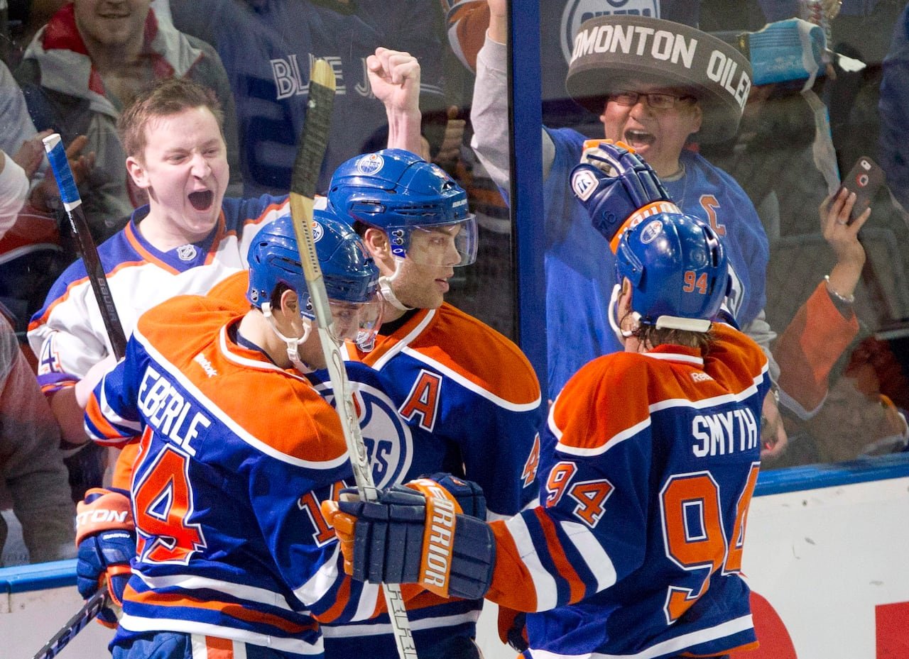 Three hockey players in orange-and-blue jerseys hug and celebrate while fans on the other side of the arena glass cheer wildly. 
