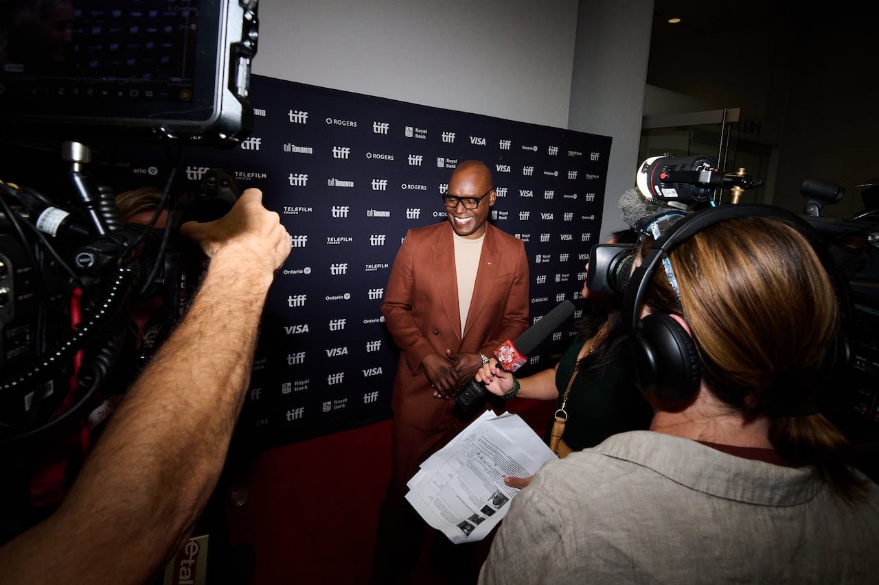 default-349 A man wearing a brown suit and glasses stands on the TIFF red carpet with cameras around him, with a black screen background behind him bearing the words "TIFF."