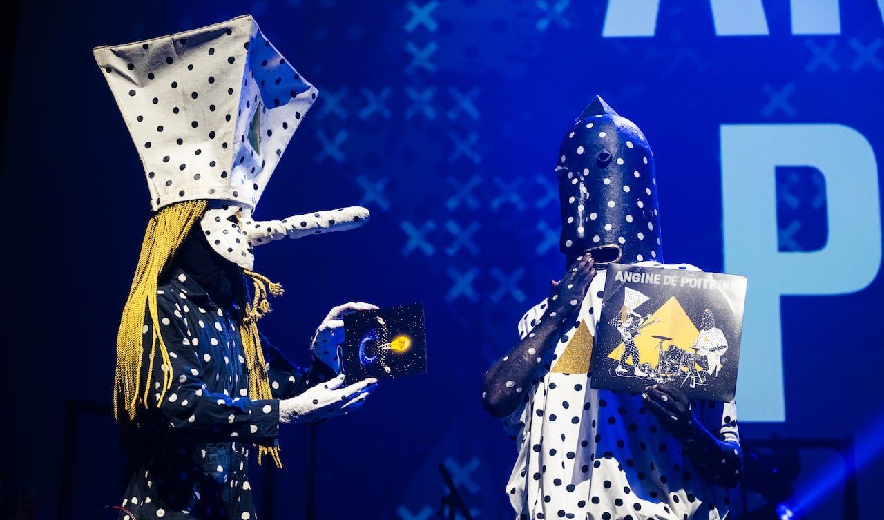 Two individuals in elaborate black-and-white polka-dotted costumes against a blue backdrop.